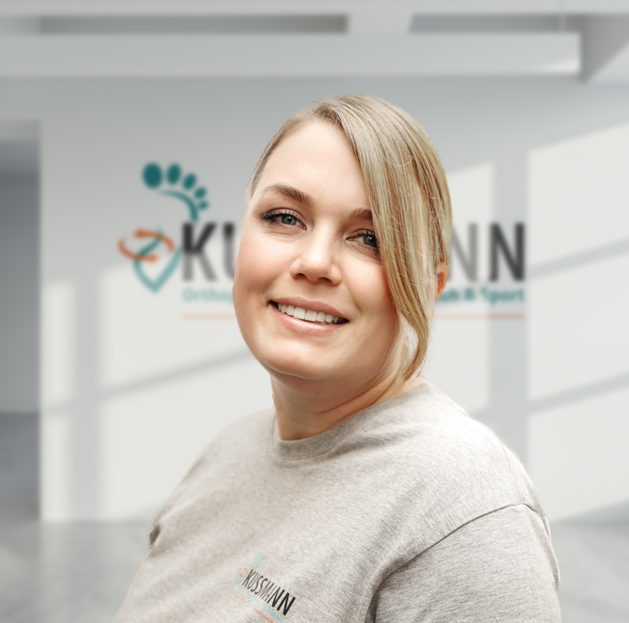 Porträt einer lächelnden Frau in einem grauen T-Shirt mit dem Logo "Kussmann" im Hintergrund. Helle, moderne Umgebung.