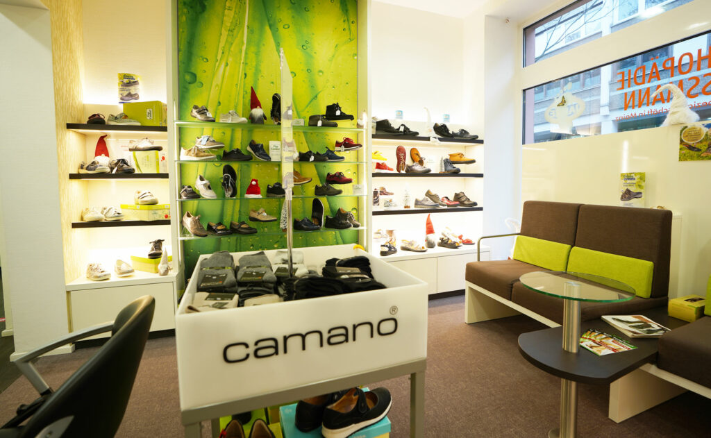 Schuhgeschäft mit Regalen voller Schuhe, einer Sitzecke und einem Tisch. Camano-Logo sichtbar. Grüner Hintergrund.