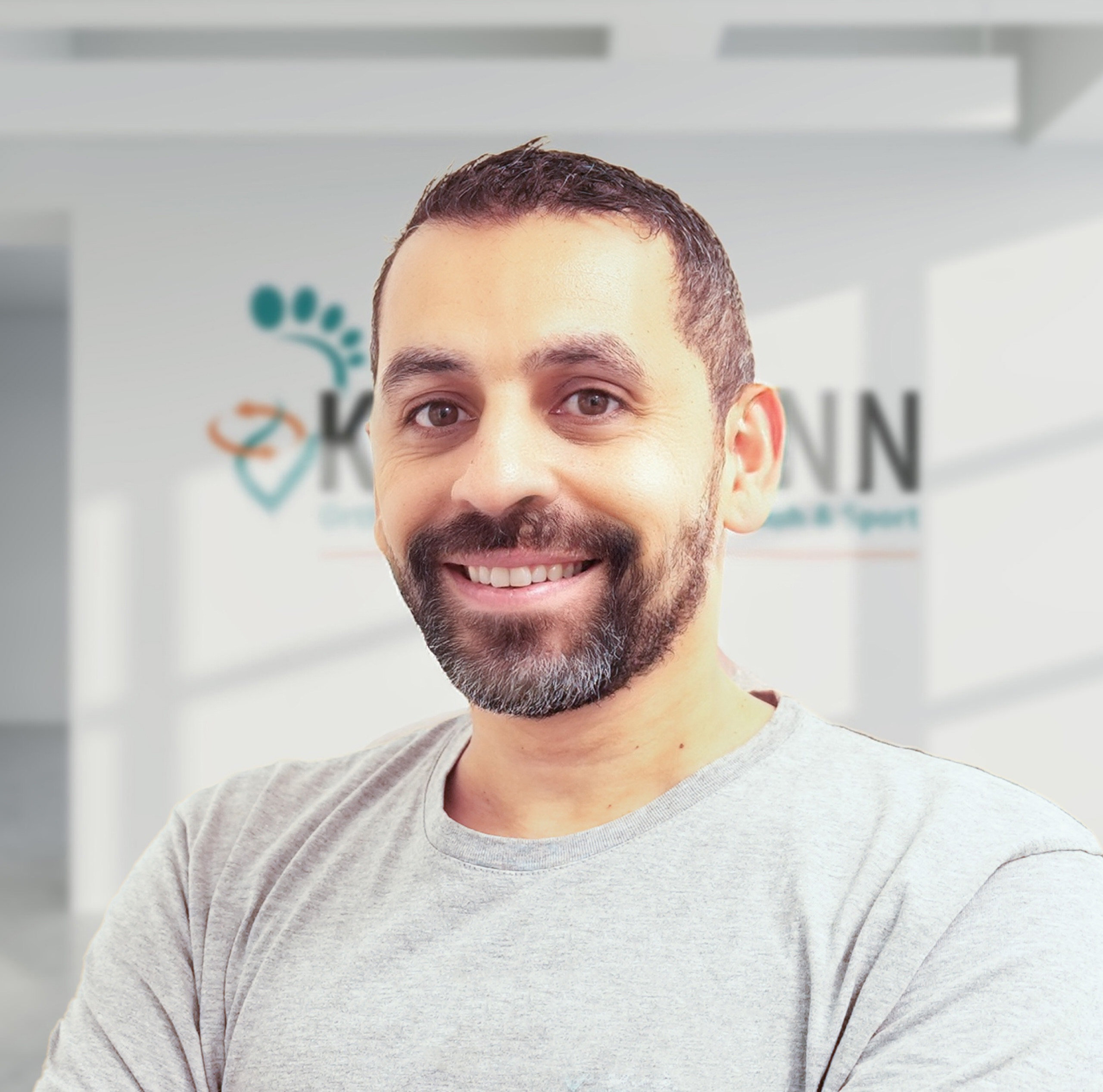 Lächelnder Mann mit Bart in grauem T-Shirt, Hintergrund mit Logo von „KANN Gesundheit & Sport“.