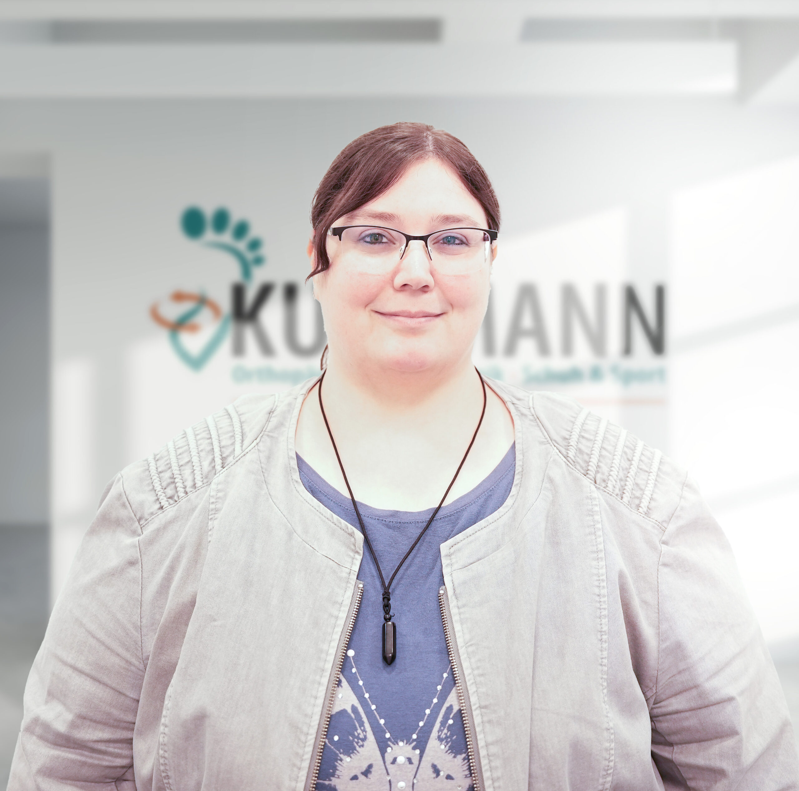 Frau mit Brille und braunen Haaren trägt eine graue Jacke und steht vor einem Hintergrund mit dem Logo „Kutzmann“.